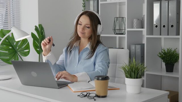 Young professional woman wearing headphones works on her laptop in stylish home office with natural light, greenery. She multitasks with coffee, documents, digital devices for remote productivity