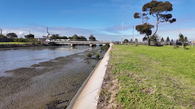 Walking perspective along the grass bank of Cherry Creek in Seaholme&nbsp;, Melbourne, Australia. Concrete lining and exposed mudbanks of the tidal waterway at low tide, leading toward road crossing.