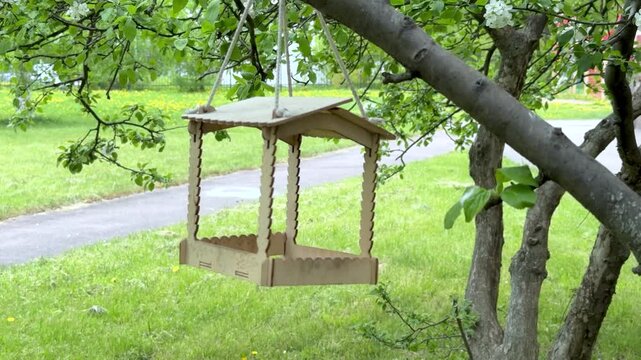 Handmade wooden bird feeder with pitched roof hangs from tree branch. Natural plywood construction features decorative cutouts. Lush green garden and foliage create serene outdoor setting. 4K footage.