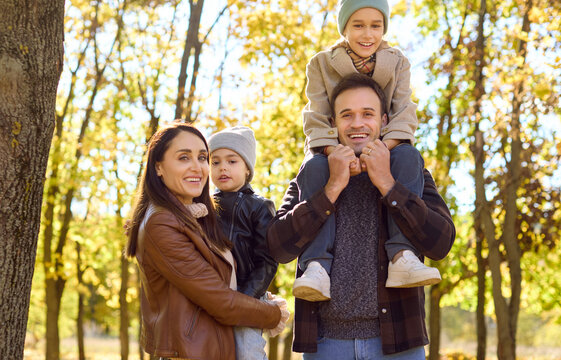 Family in autumn park, parents and children smiling. Father carries his son on shoulders while mother holds their daughter among golden trees and warm light. Concept: family unity in autumn.