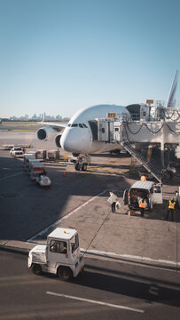 A wide-body passenger airplane parked at an airport jet bridge, surrounded by ground crew and service vehicles loading cargo under a clear sky.
