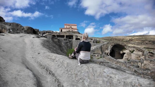 Woman sitting on ancient rock formations, enjoying historic Uplistsikhe cave city and its unique architecture carved into the cliffside, a significant cultural heritage site in Georgia near Gori city
