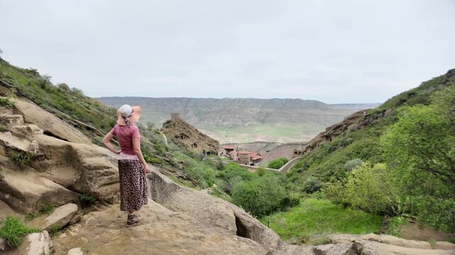 Woman resting on ancient rock steps, enjoying the panoramic view of the historic David Gareji rock-hewn monastery complex nestled in the green valley landscape of Georgia's Middle Ages heritage site