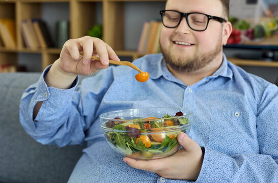 Overweight man at home eating healthy salad. Smiling adult holds a glass bowl of greens with tomatoes on a sofa, showing commitment to diet wellness. Clean indoor setting emphasizes mindful choices. 