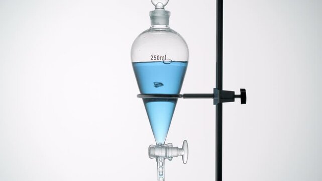 Full-frame close-up of transparent liquid pale blue draining from a separatory funnel dripping through the opened valve and causing rising bubbles inside with a light gray background