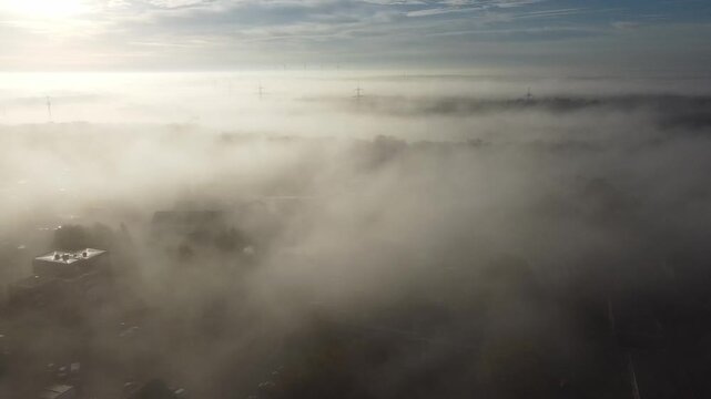 Aerial sunset landscape. Cloud fog covering misty land. Morning fog covering forest at misty sunrise. Treetop with power lines and wind turbines peek through sunset fog. Sunrise fog aerial footage
