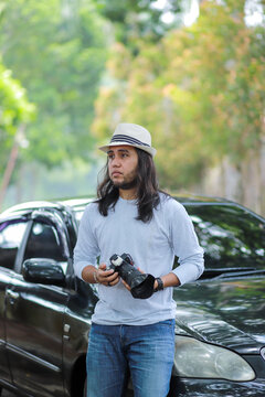 Man photographer with long hair holding camera standing beside black sedan car on tree lined street, portrait representing outdoor photography hobby and casual lifestyle concept