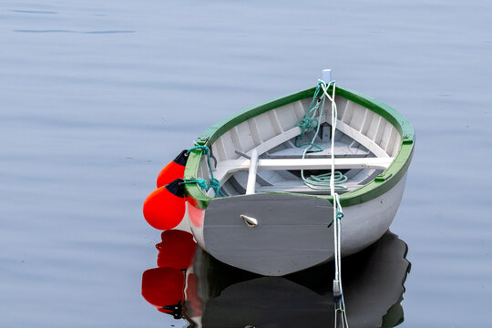A small, white colored wooden traditional dory or fishing vessel with green painted edging. The boat is floating on top of smooth water. The pond reflects the image of the recreational watercraft.