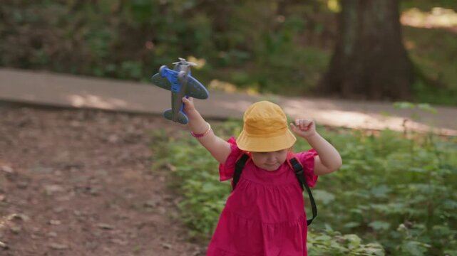 Toddler girl wearing yellow hat and pink dress running through park with toy airplane. Little child playing pilot in nature. Concept of active outdoor childhood.