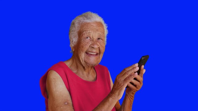 Happy senior woman using smartphone on blue chroma key background. Smiling elderly female texting and browsing on mobile phone. 4K video.