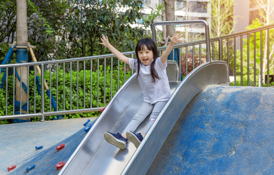 a cheerful girl sliding down a steel slide in the playground with open arms.