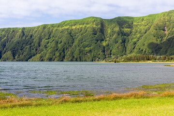 Fototapeta premium Lush green hills surround a serene lake at Sete Cidades, Sao Miguel Island, Portugal. Dense forested landscape with calm water and a clear sky. Natural volcanic environment with vibrant greenery.