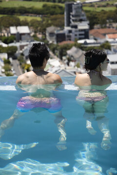 Asian couple sitting at rooftop infinity pool edge, wearing swim trunks and two-piece swimsuit