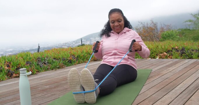 Senior African American woman pulling blue band looped around feet on mat on deck for strength