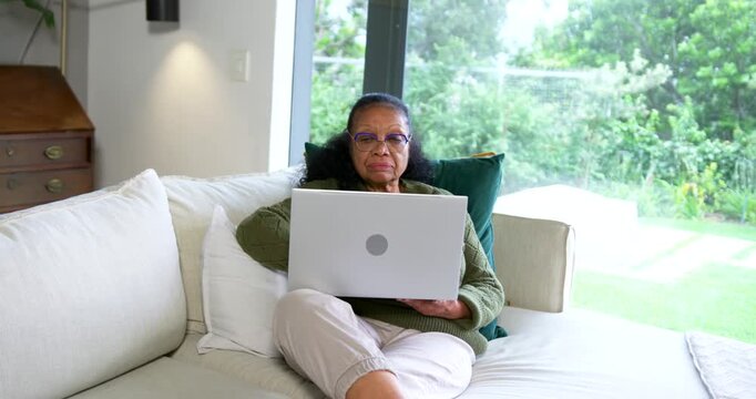 Senior African woman in green sweater placing laptop on lap on sofa, shifting, reading and typing
