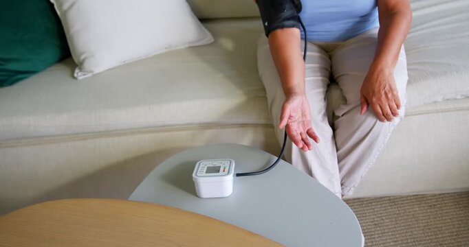 Senior woman wearing blue on sofa reaching for BP monitor, securing cuff, checking BP, copy space