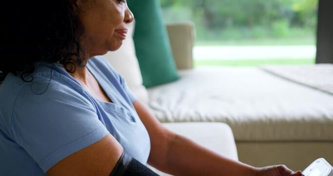Senior African American woman sitting on sofa using BP monitor cuff, pressing start for BP reading