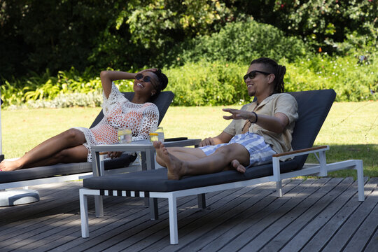 African couple reclining on gray-cushioned sun loungers, chatting on wooden deck with lemonade