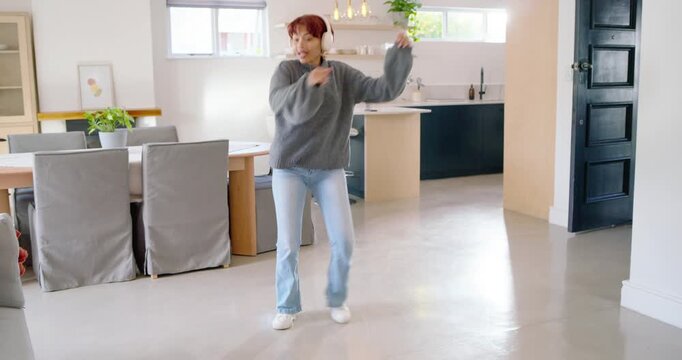 African American woman entering kitchen tapping headphones and dancing to music in gray sweater