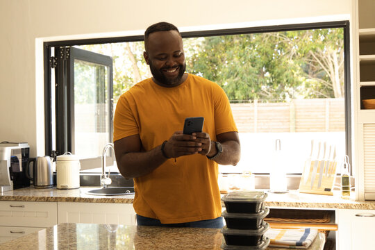African American man smiling at granite island in mustard tee holding phone stacked black prep tubs