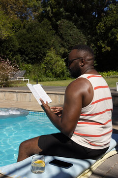 African American man in swim trunks sitting and reading pamphlet at pool with phone, lemon glass