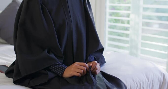 African American woman smoothing gown zipper on bed, preparing for graduation, gazing to window
