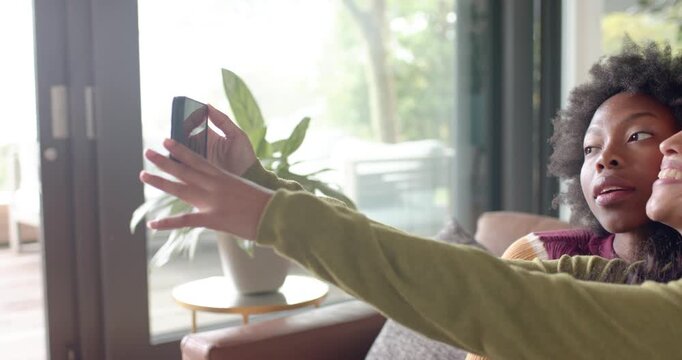 Diverse female pals leaning as mid woman extending arm holding phone while checking selfies on sofa