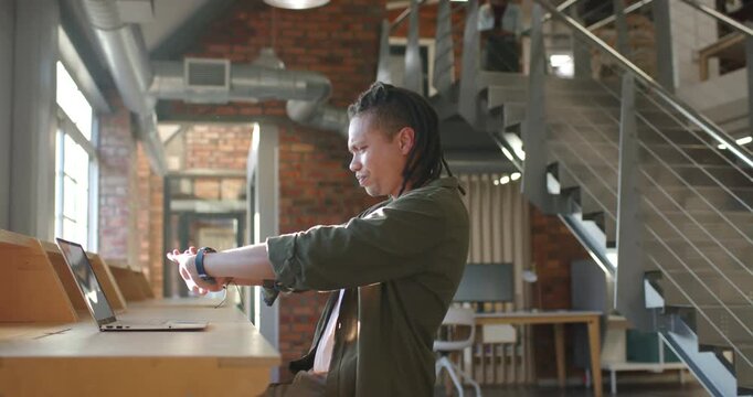 African American man in green jacket checking smartwatch alert, typing on laptop at loft for work