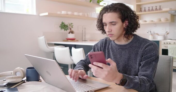 African American man in gray sweater studying red phone, typing on laptop in kitchen while working