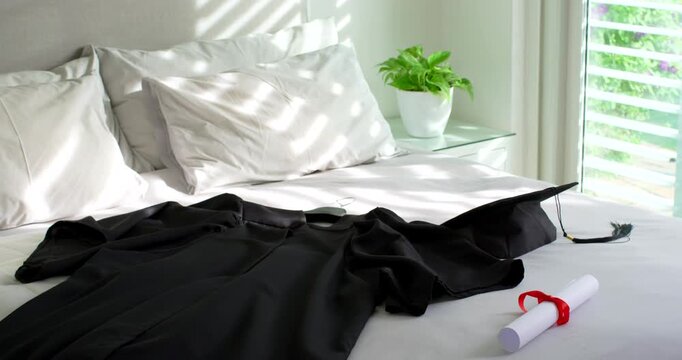 Sunlight through blinds is casting stripes across black gown, cap, diploma on bed as sun moves