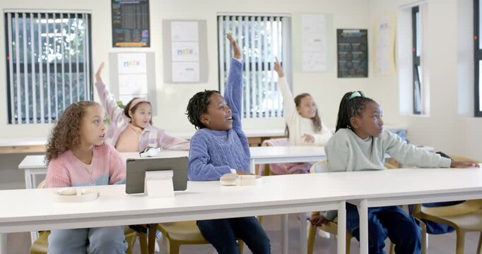 African American girls responding to offscreen teacher prompt, raising hands at classroom tablet
