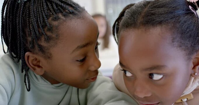 African American youth girls whispering left girl leaning in sharing secret in classroom near phone