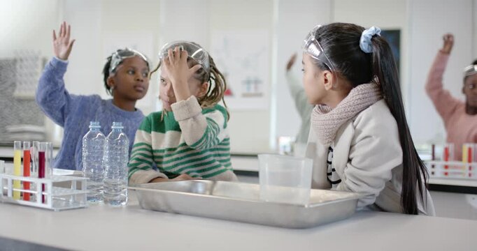 Diverse classmates sitting at lab bench wearing goggles, raising hands after teacher cue to answer