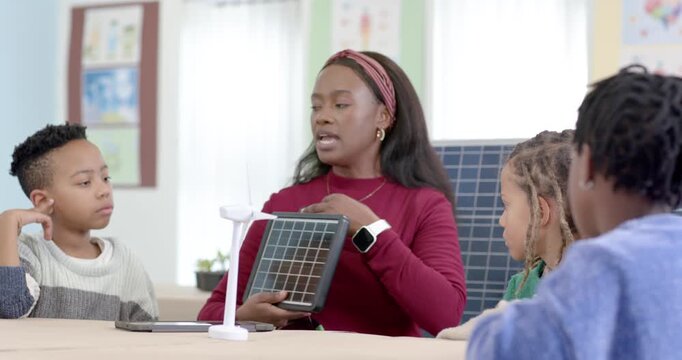 African woman teacher in maroon top teaching youth in class, picking solar panel, explaining energy