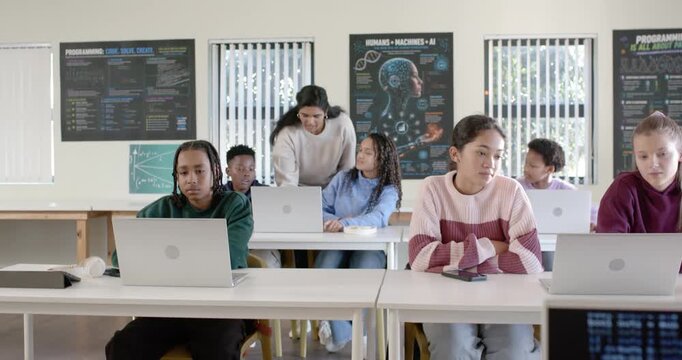 Diverse teens working on laptops in classroom as Indian teacher walking leaning in helping peer