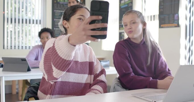 Diverse teen female friends scrolling phone, sharing screen, posing and tapping selfie in classroom