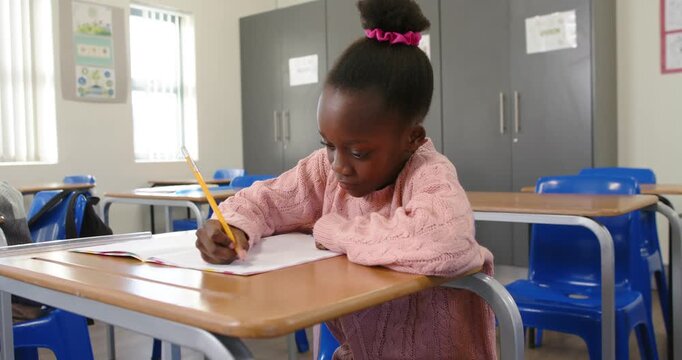 Classmates at desks, child wearing pink sweater leaning and writing in notebook with yellow pencil