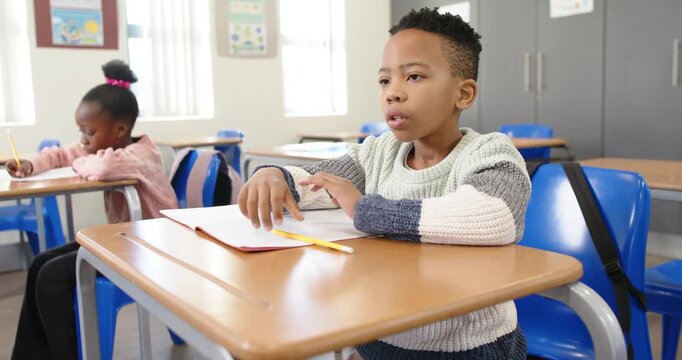 African classmates, male child writing at desk with yellow pencil, pausing, thinking, resting head
