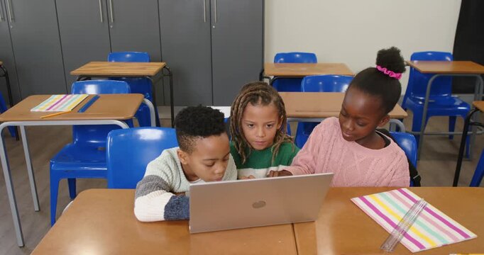 African American teens leaning and pointing at silver laptop in classroom, working on assignment