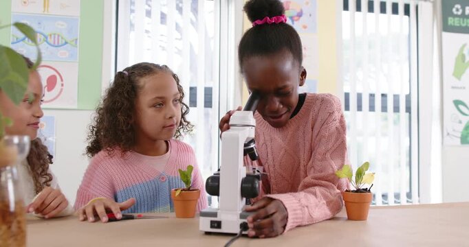 Schoolgirls watching African American girl peering into microscope inspecting plant in classroom