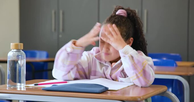 Elementary school girl looking at papers, picking up pink pencil, working on worksheet at desk