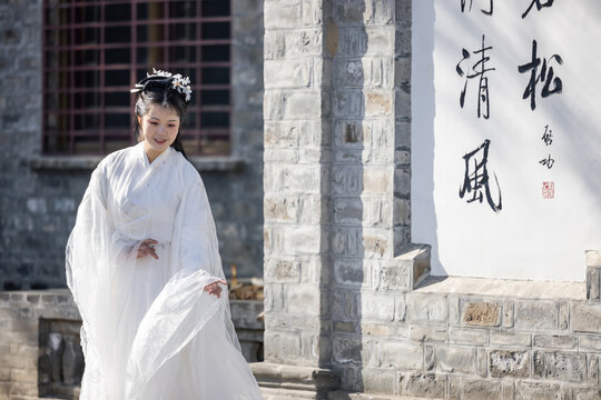 A young asian woman in white traditional hanfu poses gently outdoors beside a stone wall with chinese calligraphy, smiling softly.