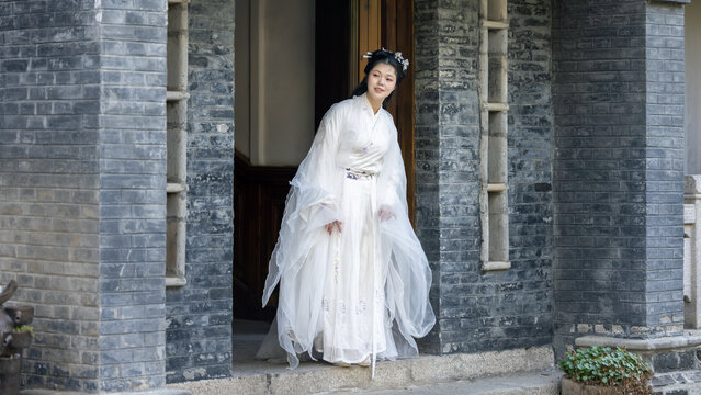 A young woman in white traditional hanfu steps out of a gray brick ancient building doorway, appearing graceful.