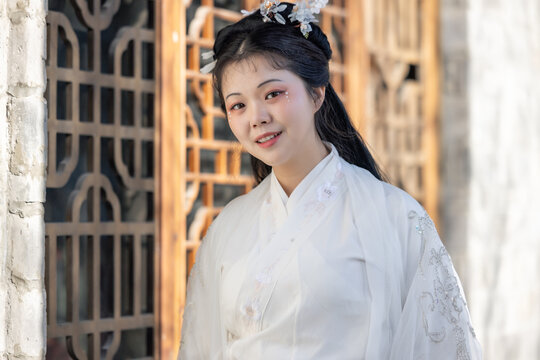 A young east asian woman wearing white traditional hanfu, with an elegant hairstyle and hairpin, smiling while standing beside a wooden lattice window of a traditional building.