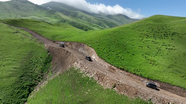 Drone shot of off-road buggies driving along a dirt path through picturesque green mountain hills. Cinematic landscape featuring rolling alpine meadows and scenic mountain ranges