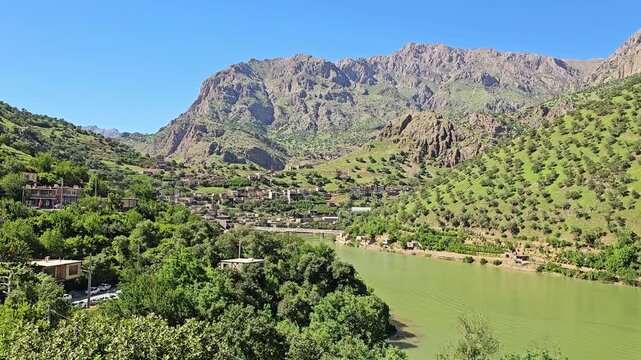 Tranquil green river winds beside a lush hillside village, framed by rocky spring peaks and orchard-covered slopes under a clear blue sky.
📍Selin village, Hawraman Takht, Kurdistan, Iran 