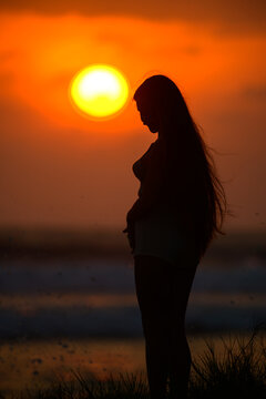 Silhouette of a woman standing at sunset by the ocean with a glowing orange sky