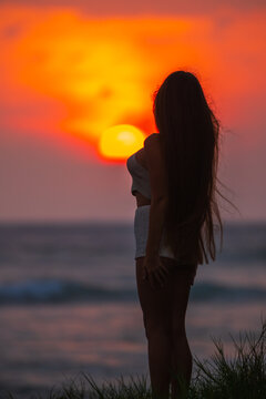 Silhouette of a woman standing at sunset by the ocean with a glowing orange sky