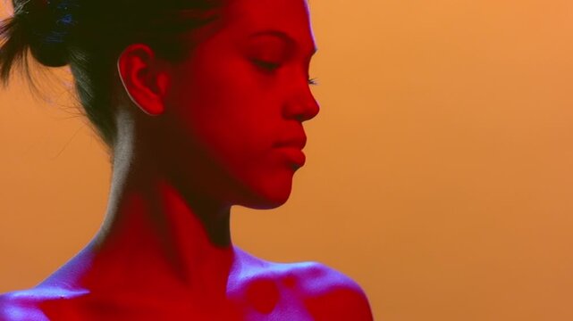 Portrait close-up of young natural beauty model illuminated with red and blue turning her head and looking aside on an orange background | Perfume advertising footage