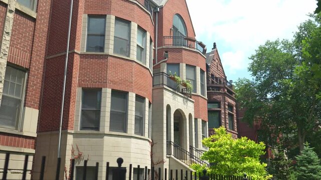 Large historic brick residential building with balcony, arched entrance, and decorative architectural details. Sunlight highlights the facade and greenery in a quiet urban neighborhood.